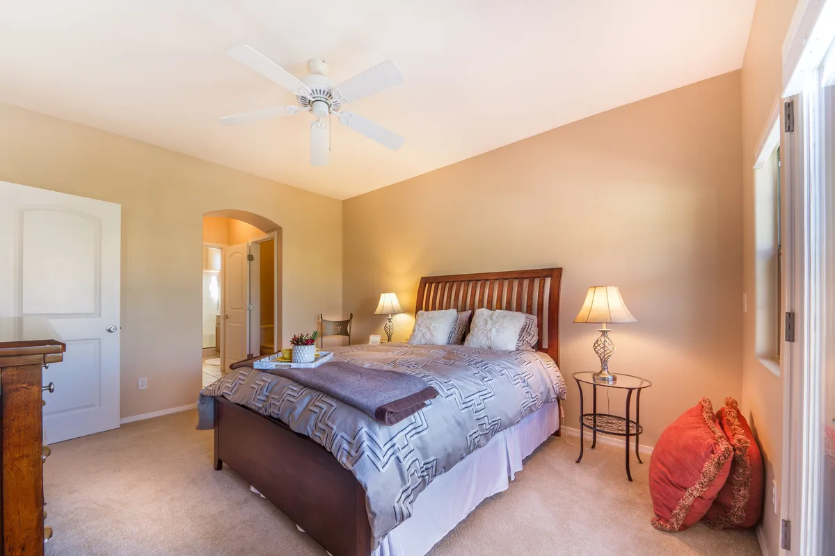 A serene bedroom featuring a queen bed adorned with decorative pillows and a soft blanket. Bedside lamps create a warm glow, while a wooden dresser completes the cozy, inviting look. Natural light streams in from a window, enhancing a tranquil mood—ideal for restful nights and peaceful mornings.