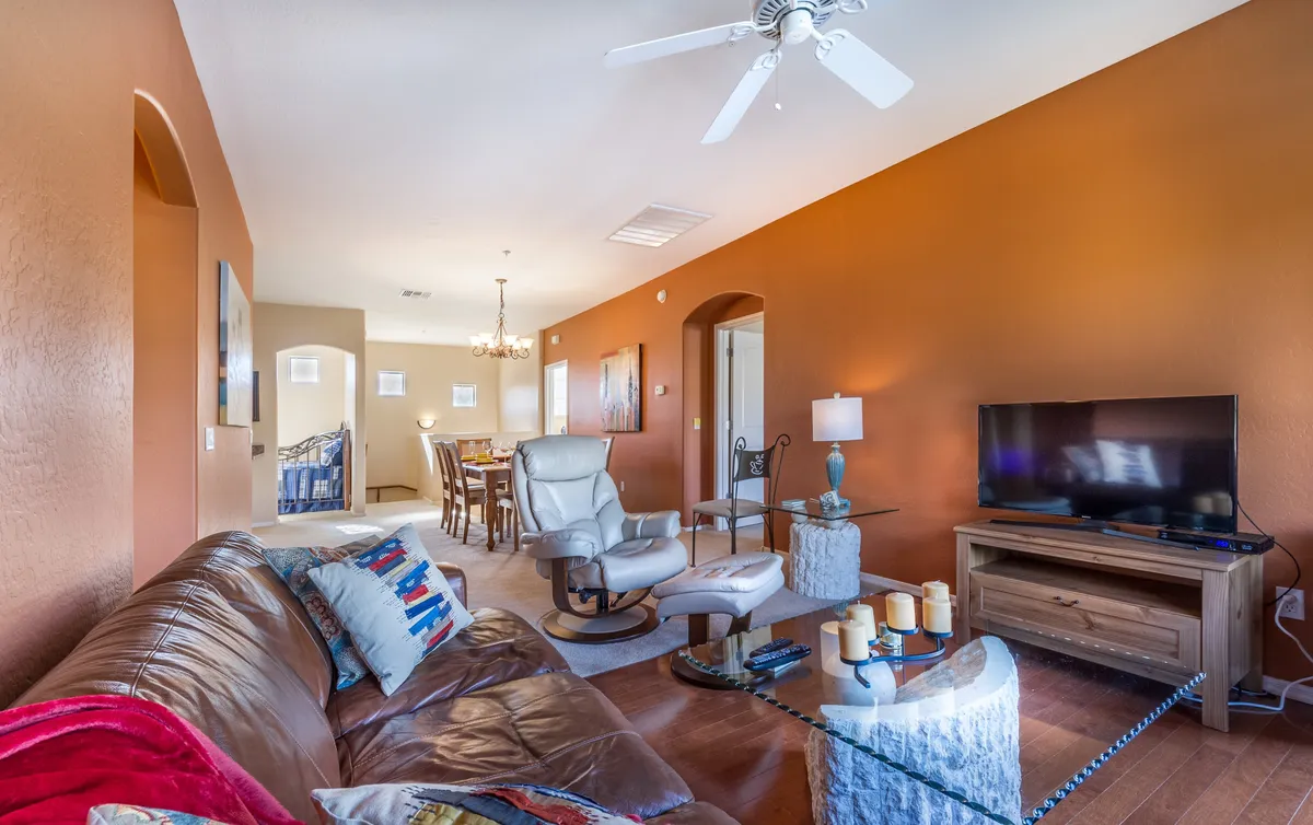 A cozy living room with a brown leather sofa, glass coffee table, TV stand with television, and a dining area visible in the background. Natural light streams in, creating a comfortable and inviting space suited for both relaxation and entertainment.