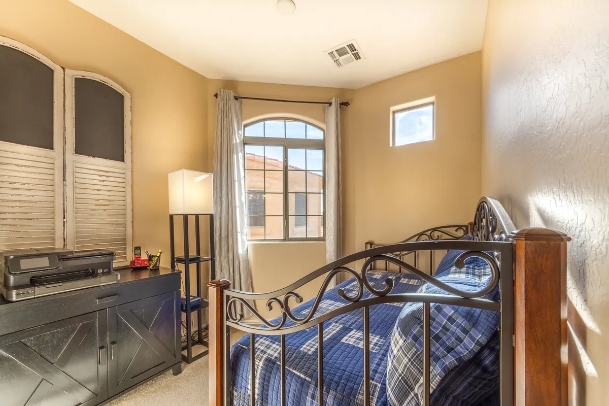 A serene bedroom scene with a comfortable bed, large window letting in natural light, and a lamp atop wooden furniture. Notably, the room features a desk, ideal for work sessions or creative pursuits. The setup embodies the ideal remote work environment—peaceful, bright, and functional.
