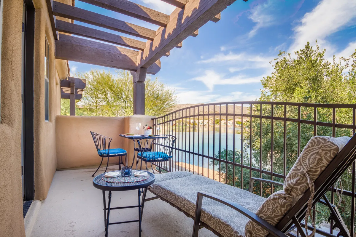 An exterior view of a bistro table with chairs and a lounge chair set on a patio overlooking a serene lake. The scene is tranquil, with morning sunlight reflecting on the water, inviting relaxation, outdoor dining, and social gatherings in a picturesque setting.