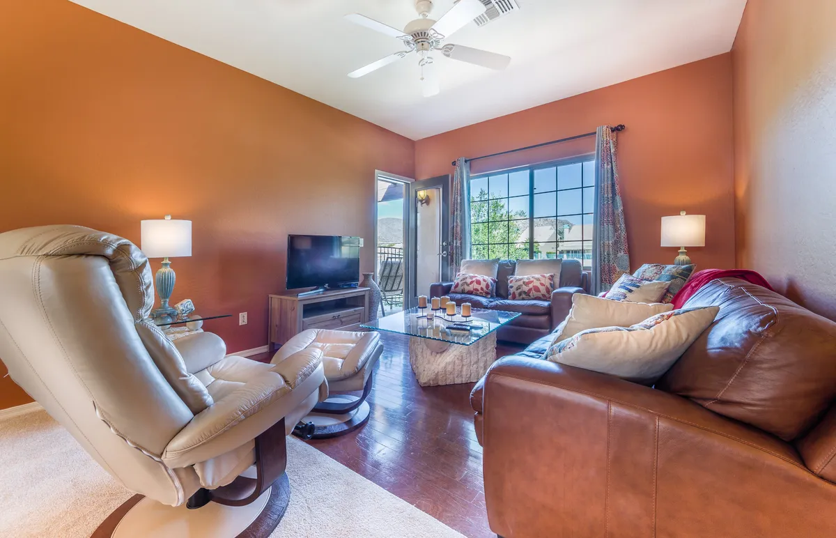 The inviting living area features a large window streaming in natural light, a leather couch, a coffee table, and a TV. The setting is perfect for relaxing, watching a movie, or having intimate conversations, bathed in afternoon sunlight.