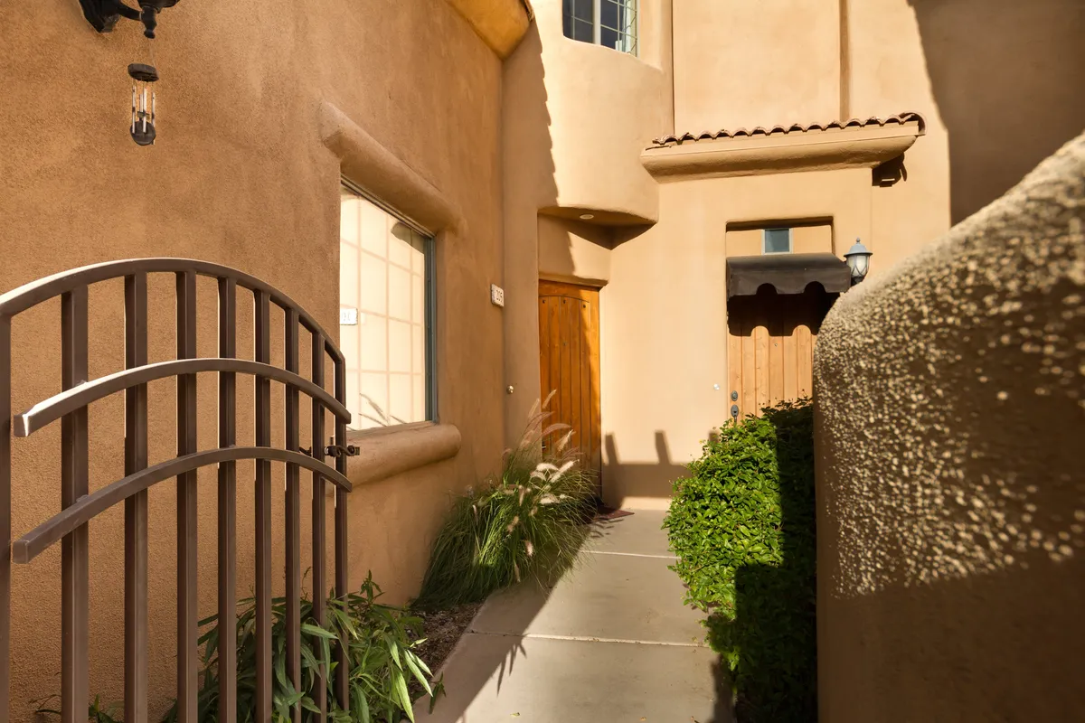 A charming arched metal gate leads to a wooden front door surrounded by lush greenery and a textured wall, emphasizing the gated community's secure and private entrance. The pathway suggests a peaceful, welcoming environment for residents and guests alike.