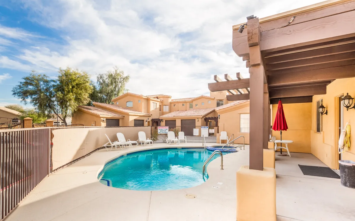 This image features a sparkling swimming pool surrounded by lounge chairs and a vibrant red umbrella, with a pergola providing shaded seating. The scene exudes a relaxed, resort-like atmosphere, perfect for socializing, swimming, or sunbathing. The pool is framed by well-maintained landscaping, conveying a sense of privacy and exclusivity within the community.