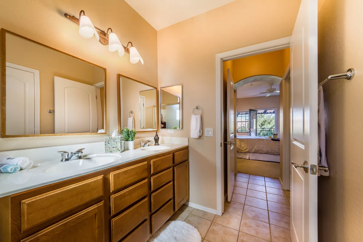 A bright, spa-inspired bathroom with a double sink vanity, large mirrors, elegant light fixtures, and green decorative plants. The view into the adjacent bedroom hints at seamless flow and privacy. The fresh, sophisticated design emphasizes personal space and luxurious self-care.
