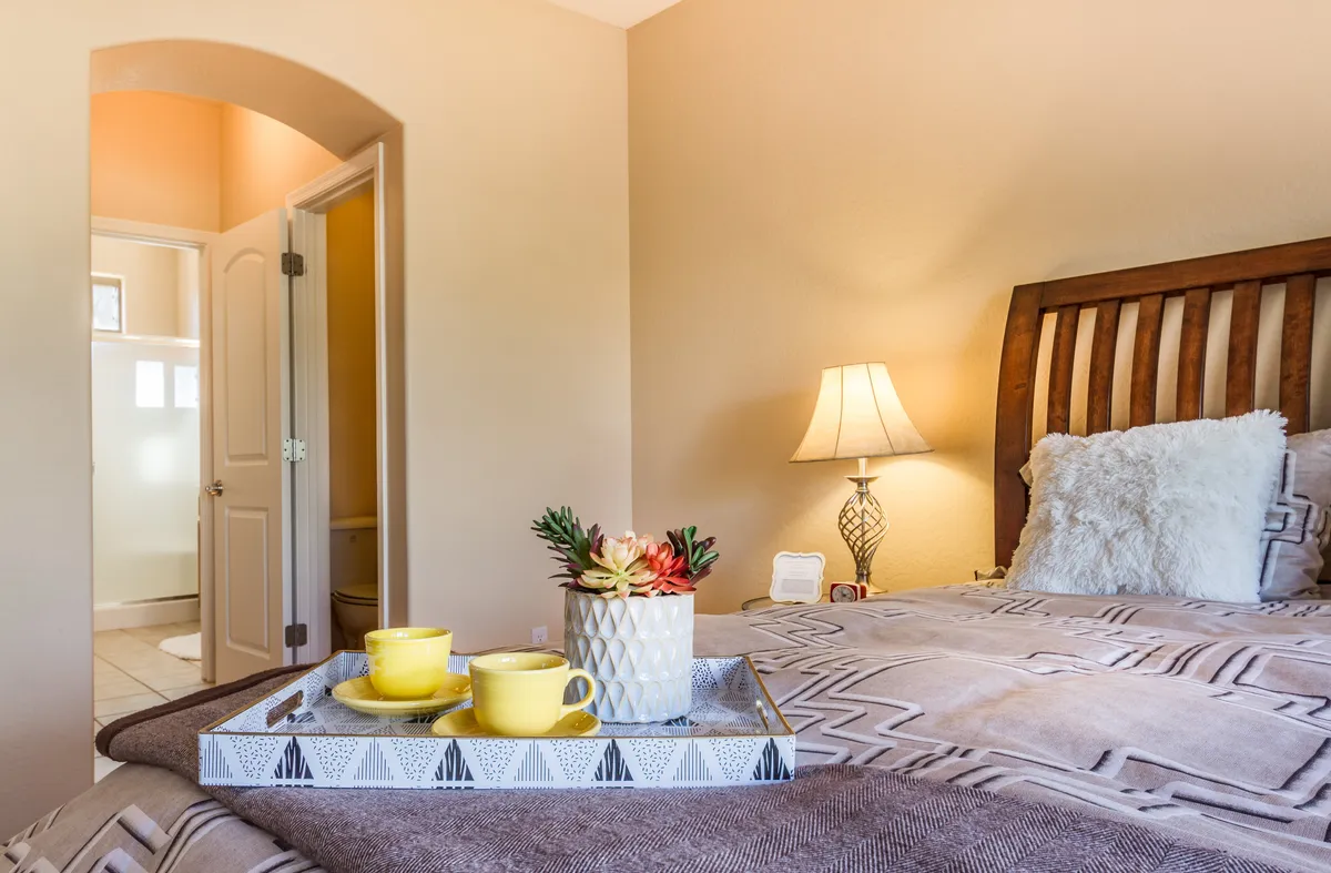 This image captures a cozy bedroom scene featuring a bed with decorative pillows, a nightstand with a lamp for soft ambient lighting, and a tray adorned with cups and a decorative plant. The warm, inviting atmosphere suggests a perfect spot for sipping morning coffee and relaxing in comfort.